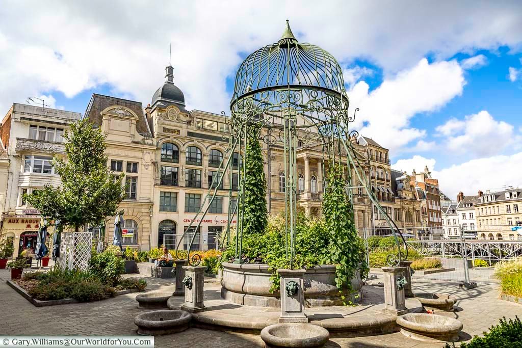 The decorative Vieux Puits fountain in Saint-Quentin is surrounded by elegant historic buildings and landscaped public spaces beneath bright summer skies. This charming landmark offers a peaceful stop during a Saint-Quentin walking tour and reflects the city’s rich architectural heritage.