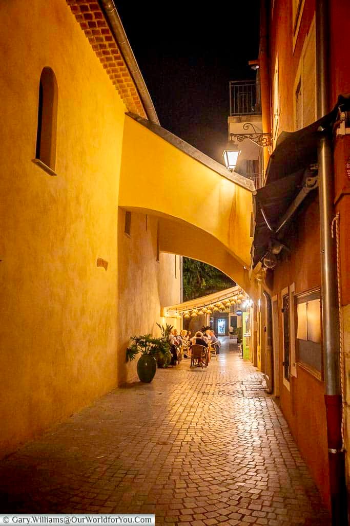 An atmospheric narrow passage in Sanary-sur-Mer after dark, where warm streetlights lead beneath an archway towards a small outdoor restaurant.