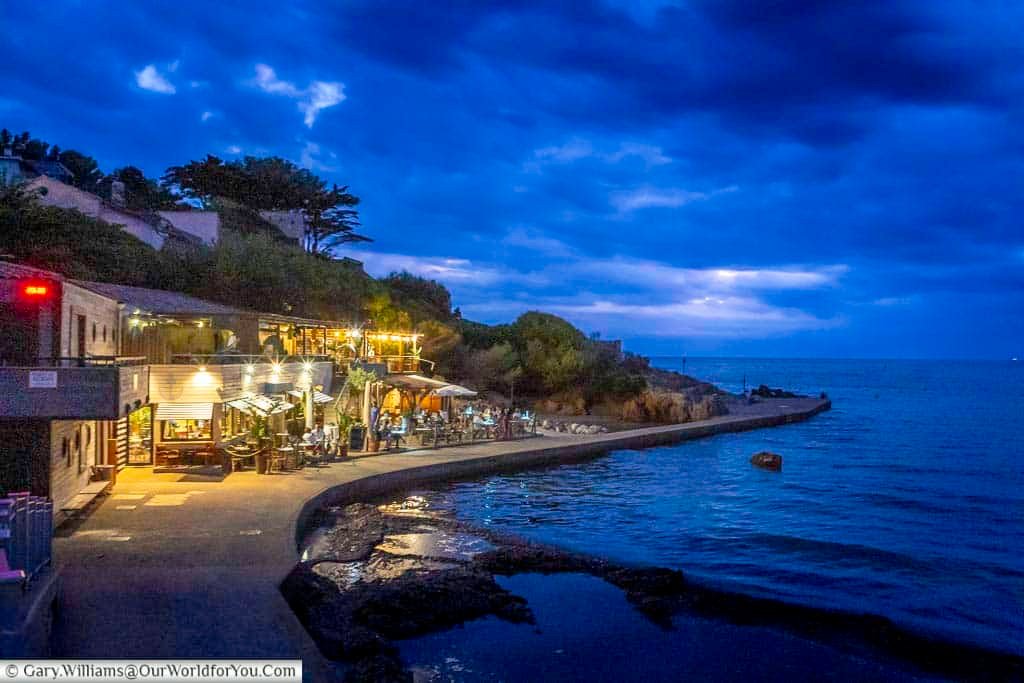 A peaceful evening beside Portissol Bay in Sanary-sur-Mer, with a warmly lit waterfront restaurant overlooking the Mediterranean beneath a dramatic blue twilight sky.