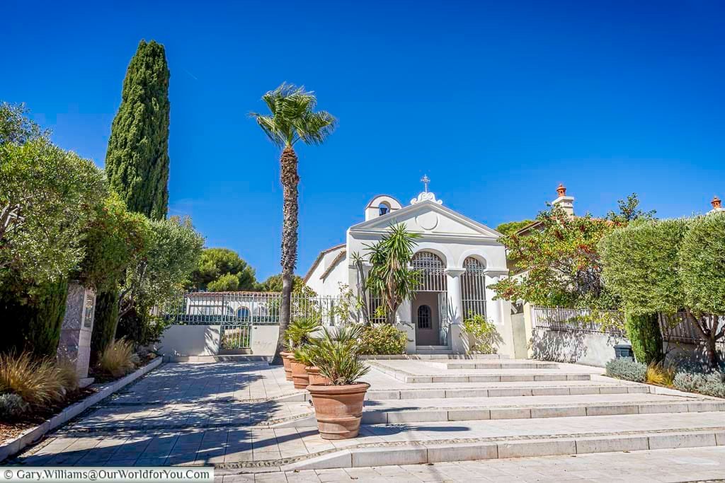 The white Chapel Notre-Dame de Pitié in Sanary-sur-Mer, surrounded by palms and Mediterranean greenery beneath a vivid blue Provençal sky.