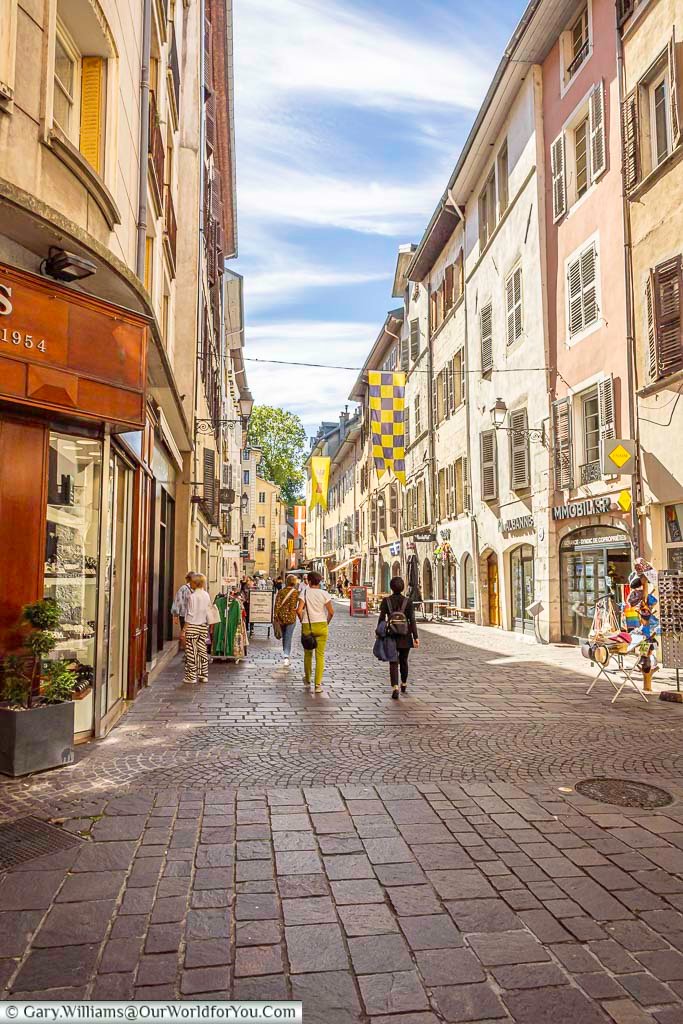A lively cobbled shopping street winds through the historic centre of Chambéry, lined with colourful Savoyard buildings, traditional shutters and local shops. Decorative banners overhead add to the character of this attractive pedestrian-friendly area of the old town.