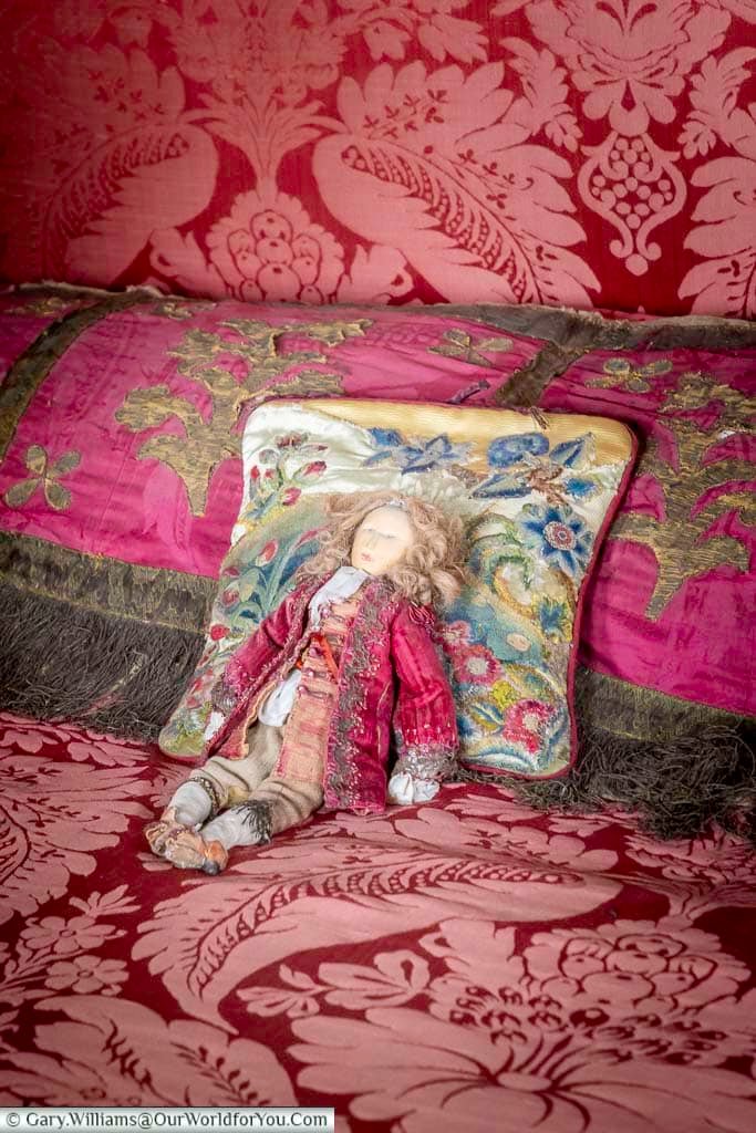 Historic-style doll resting among the richly coloured textiles and embroidered cushions of the King's Bed Chamber at Restoration House in Rochester.