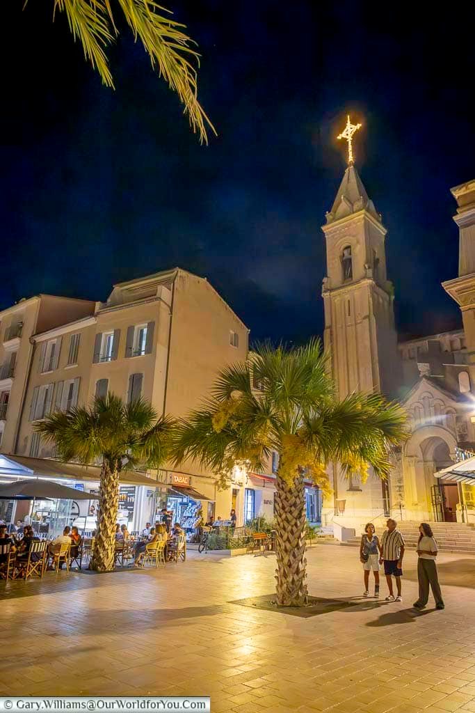The bell tower of Saint-Nazaire Church rising above the palm-lined streets and lively café terraces of Sanary-sur-Mer after dark.