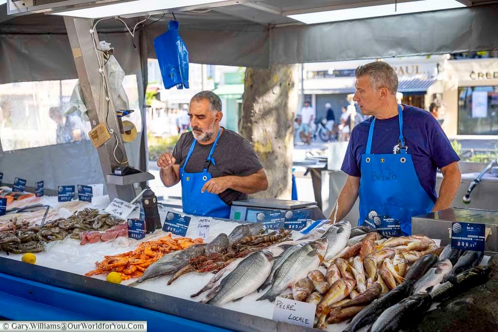 Local fishmongers behind a colourful seafood stall at Sanary-sur-Mer market, with freshly caught fish, prawns and shellfish displayed on ice.