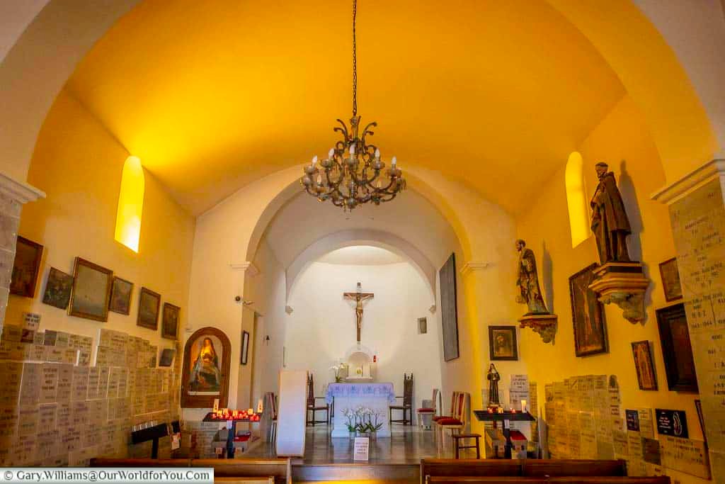 The intimate interior of Notre-Dame de Pitié in Sanary-sur-Mer, with its simple altar, religious statues, votive plaques and warm golden light.