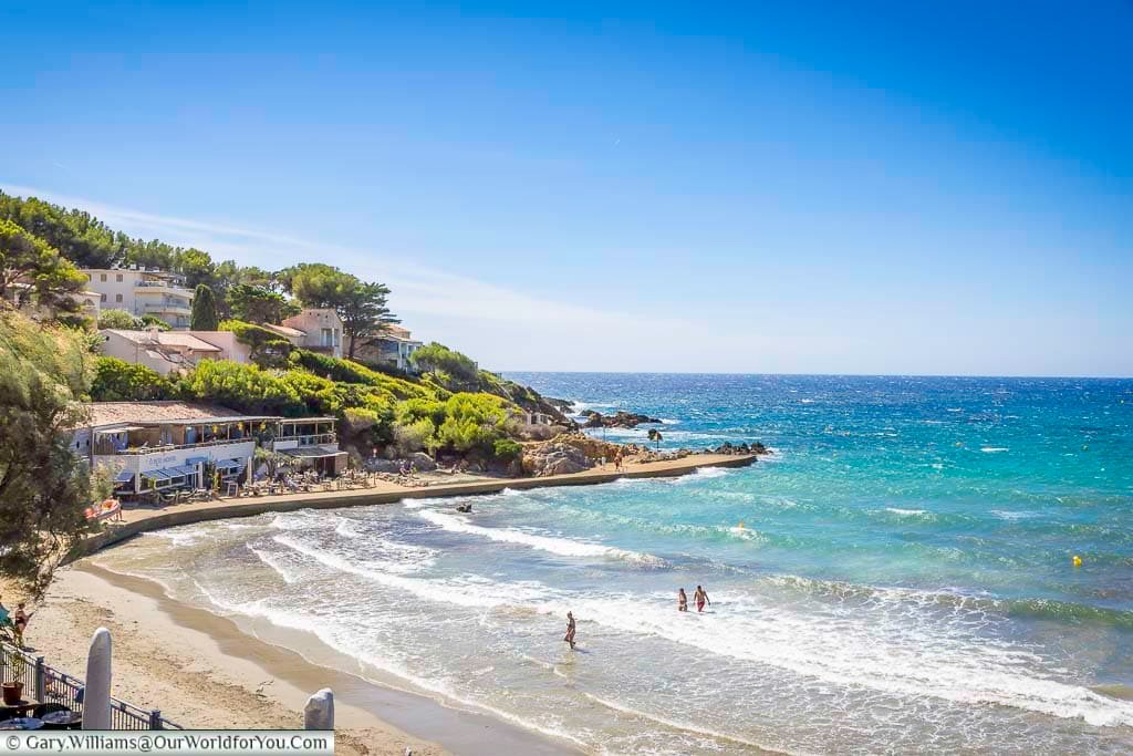 The beautiful sheltered bay of Portissol Beach in Sanary-sur-Mer, with turquoise Mediterranean water, a sandy beach and rocky pine-covered shoreline.