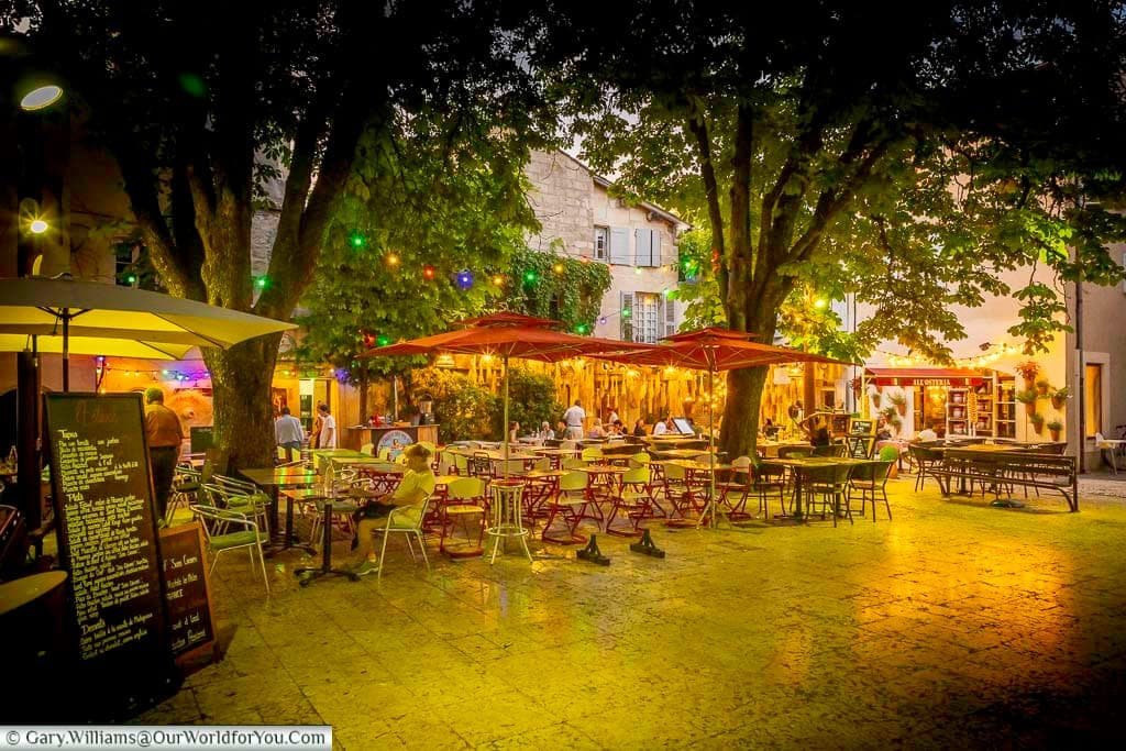 Atmospheric outdoor restaurants beneath leafy trees and colourful lights on a summer evening in Saint-Rémy-de-Provence, France.