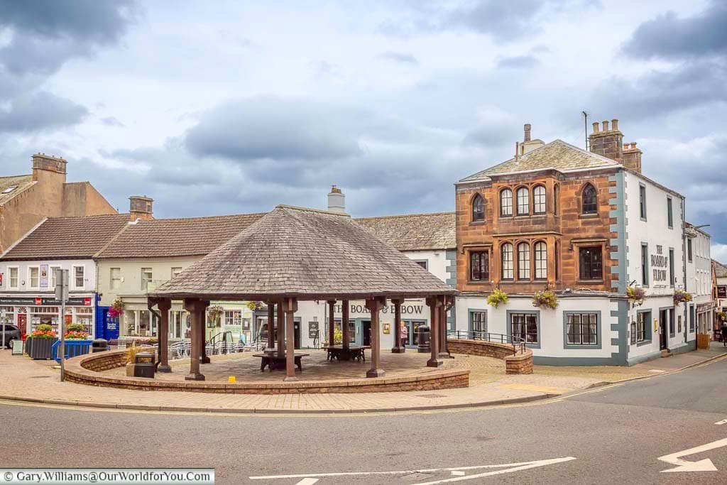 A traditional timber-roofed shelter stands in a picturesque square in Penrith, surrounded by historic buildings and local shops. This charming corner of the Cumbrian market town reflects its welcoming atmosphere and architectural heritage.