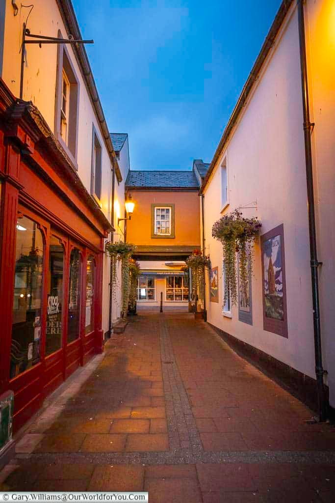 A narrow historic pedestrian lane in Penrith is lined with traditional buildings, independent shops and colourful hanging flower baskets. Warm evening lighting enhances the charm of this picturesque corner of the Cumbrian market town.
